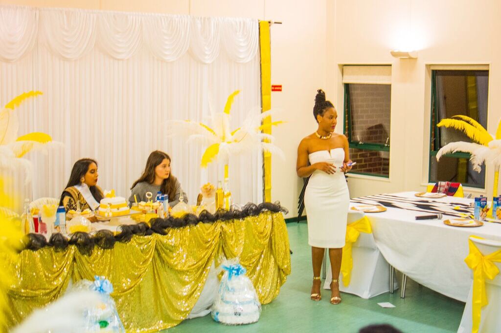 A woman in a white dress stands beside a decorated table, addressing three seated women. The room features gold and white decorations with feathered centerpieces and wrapped gift baskets. A woman in a white dress stands beside a decorated table, addressing three seated women. The room features gold and white decorations with feathered centerpieces and wrapped gift baskets.