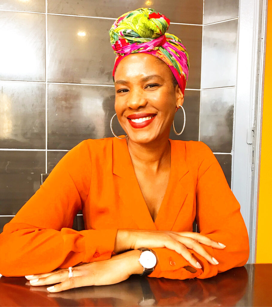 ca13b9cd-9c67-4997-856c-84f2cd7d32c1 A woman wearing a vibrant orange blouse and a colorful headscarf smiles at the camera. She is sitting with her hands on a table, wearing large hoop earrings and a watch. The background features dark, reflective tiles.