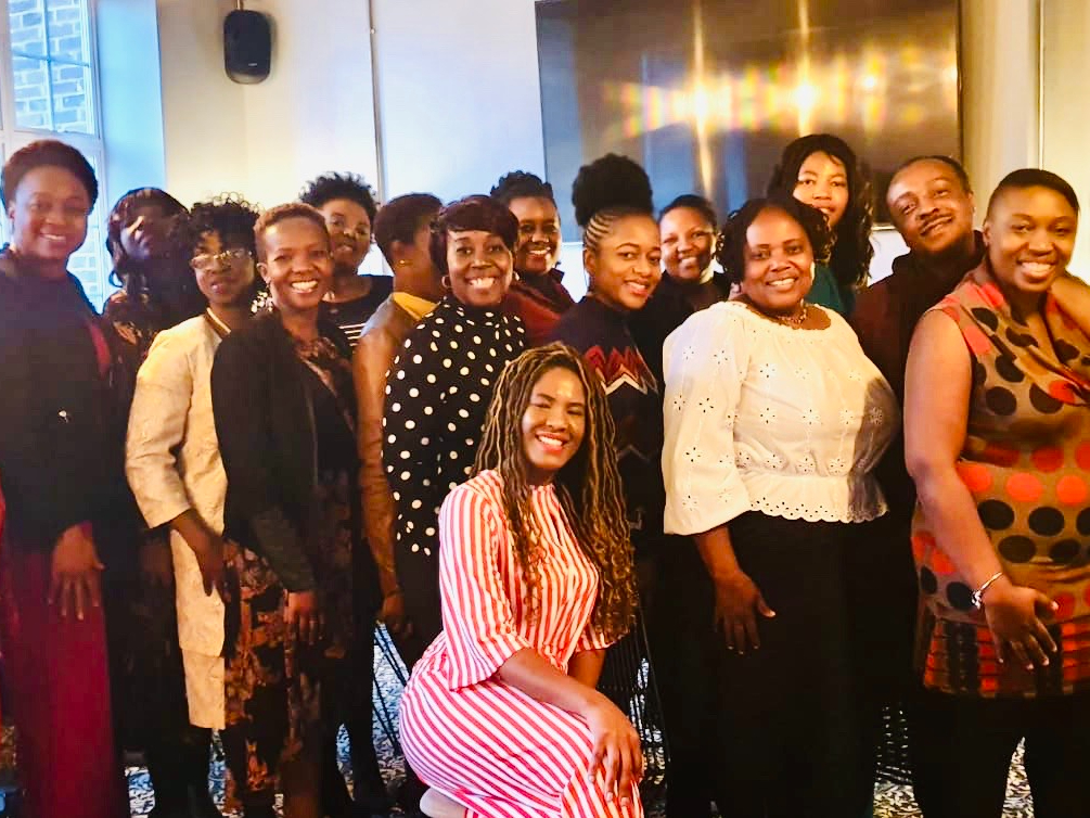 A group of people smiling and posing together indoors, radiating camaraderie. One person sits in the front wearing a red and white striped outfit, while the others stand behind, dressed in various outfits. Their shared bond reflects principles of intentional parenting coaching seen across the UK. A group of people posing together indoors, smiling at the camera. One person in the front is seated wearing a red and white striped outfit. Others are standing, wearing a variety of colorful clothing. The background is a warmly lit room.