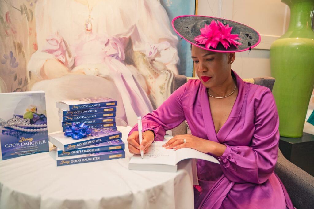A person in a vibrant purple outfit and hat signs a book at a table. A stack of books titled "Being God's Daughter" is nearby. The setting is elegant, with a large painting and a decorative vase in the background.