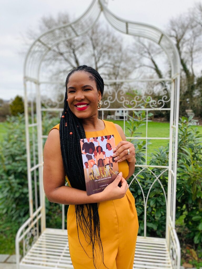 A woman in a yellow dress stands outdoors in a white gazebo, smiling and holding a book with a colorful cover. The background features greenery and trees.