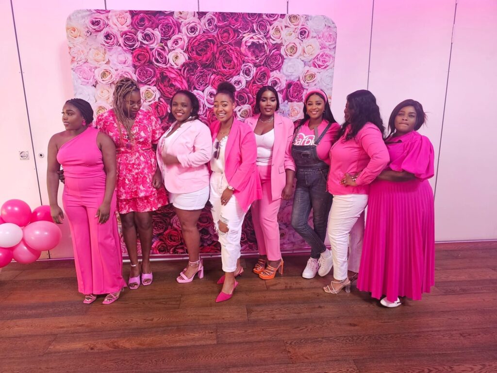 A group of eight women stands in front of a floral backdrop at an intentional parenting event in the UK, wearing various shades of pink and white outfits. They pose together, smiling. Some wear dresses, while others are in pants, with pink balloons visible on the left.