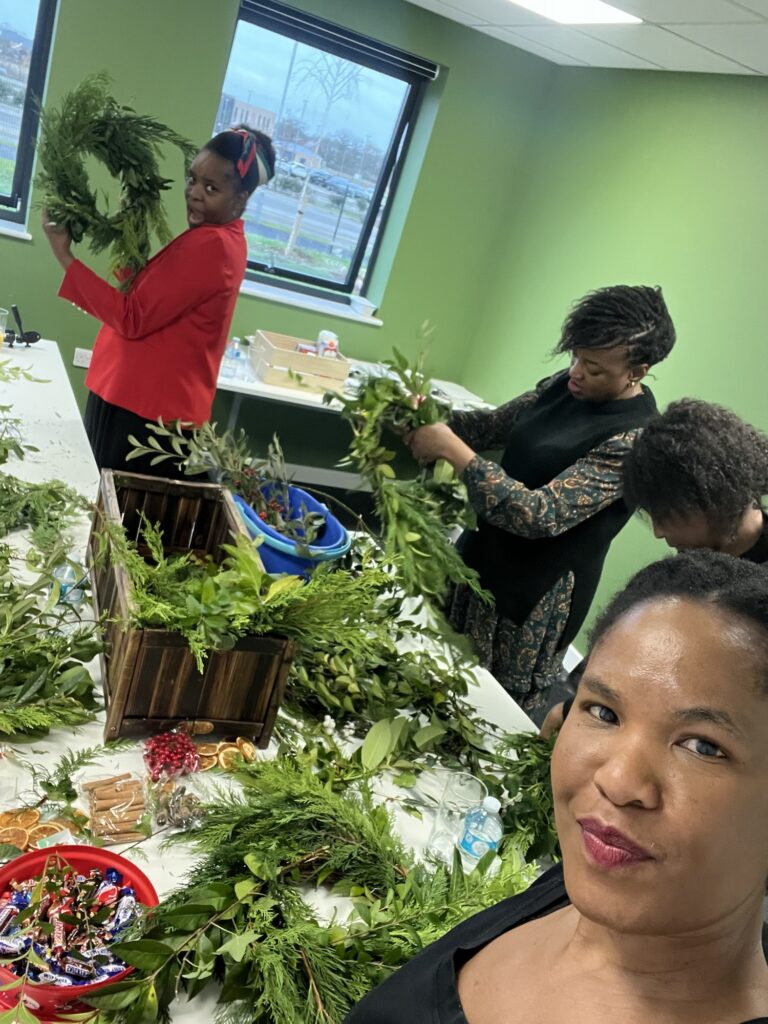 Four people are standing around tables covered in greenery and craft supplies. They are engaged in creating wreaths or arrangements. One person takes a selfie, smiling at the camera. The room has green walls and a window showing an outside view.