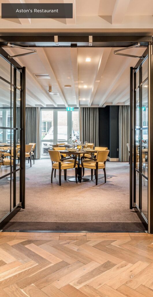 Glass doors opening to a modern restaurant with neatly arranged tables and beige chairs. Natural light streams through large windows with gray curtains, illuminating the wooden flooring. A sign above reads "Aston's Restaurant.