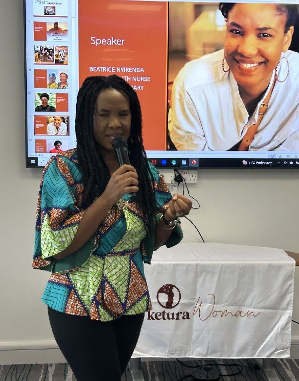 A woman with long hair speaks into a microphone in front of a presentation screen. The slide shows another woman smiling with the title "Speaker" and a logo on a tablecloth reads "Ketura Woman.