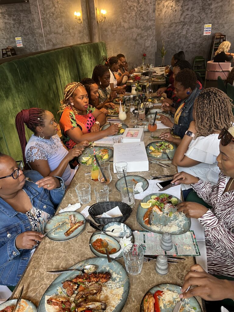 A group of people sitting at a long table in a restaurant, enjoying a meal. Plates of food with rice, meats, and salad are in front of them. Some are holding menus, and drinks are on the table. The setting is casual and lively.