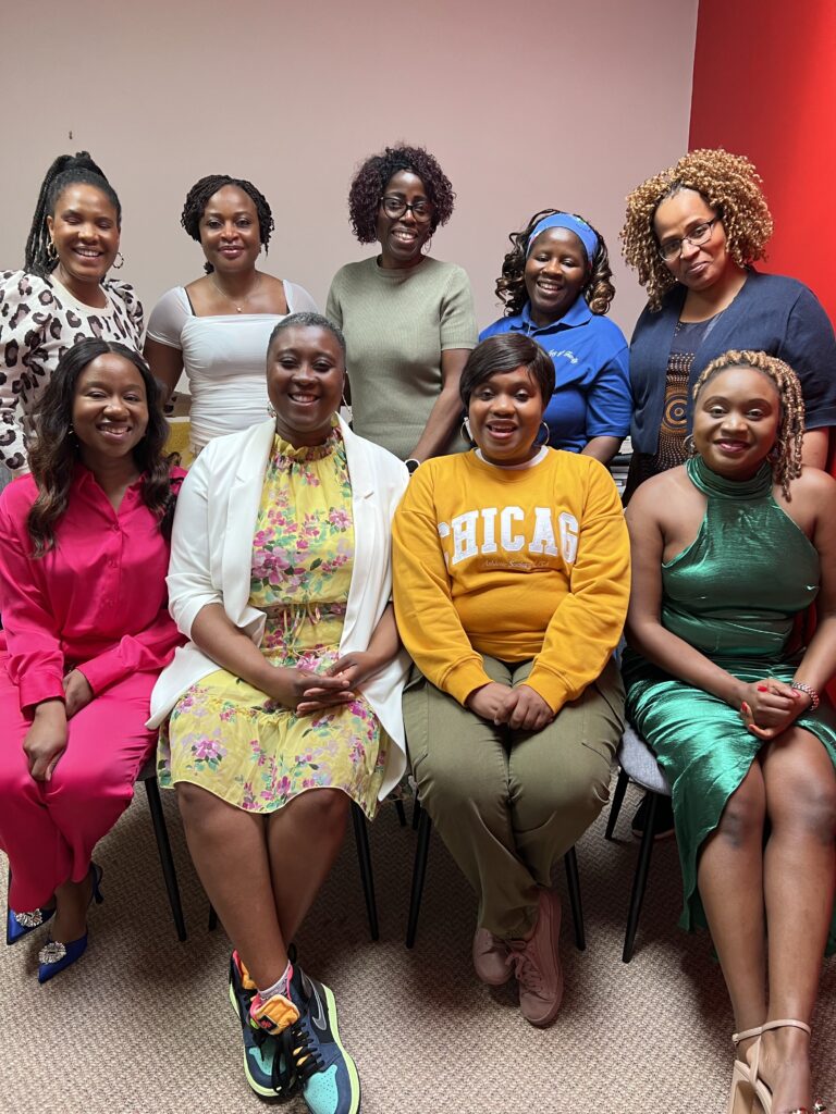 A group of nine women are posing together indoors. They are dressed in a variety of colorful and stylish outfits, and all are smiling at the camera. The setting appears to be a warmly lit room with a red and white wall.